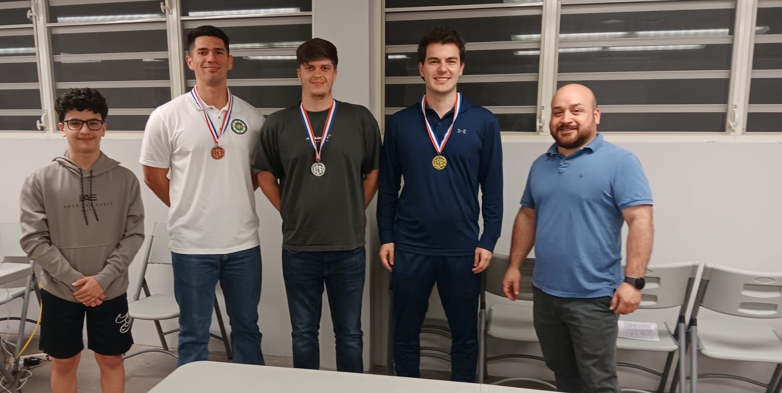 Cinco personas de pie en el Pabellón de la Escuela Luis Muñoz Rivera; tres llevan medallas de la 2da Copa Gambito del Otoao.