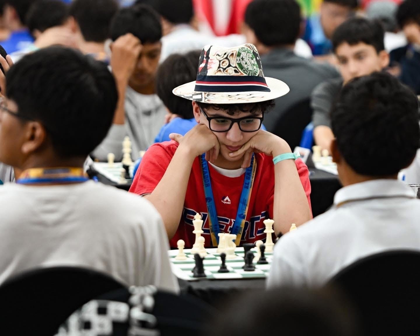 Stefan J. Marín Rivera, con sombrero y camisa roja, concentrado con la barbilla en la mano frente al tablero, rodeado de jugadores en el Campeonato Panamericano de la Juventud en El Salvador.