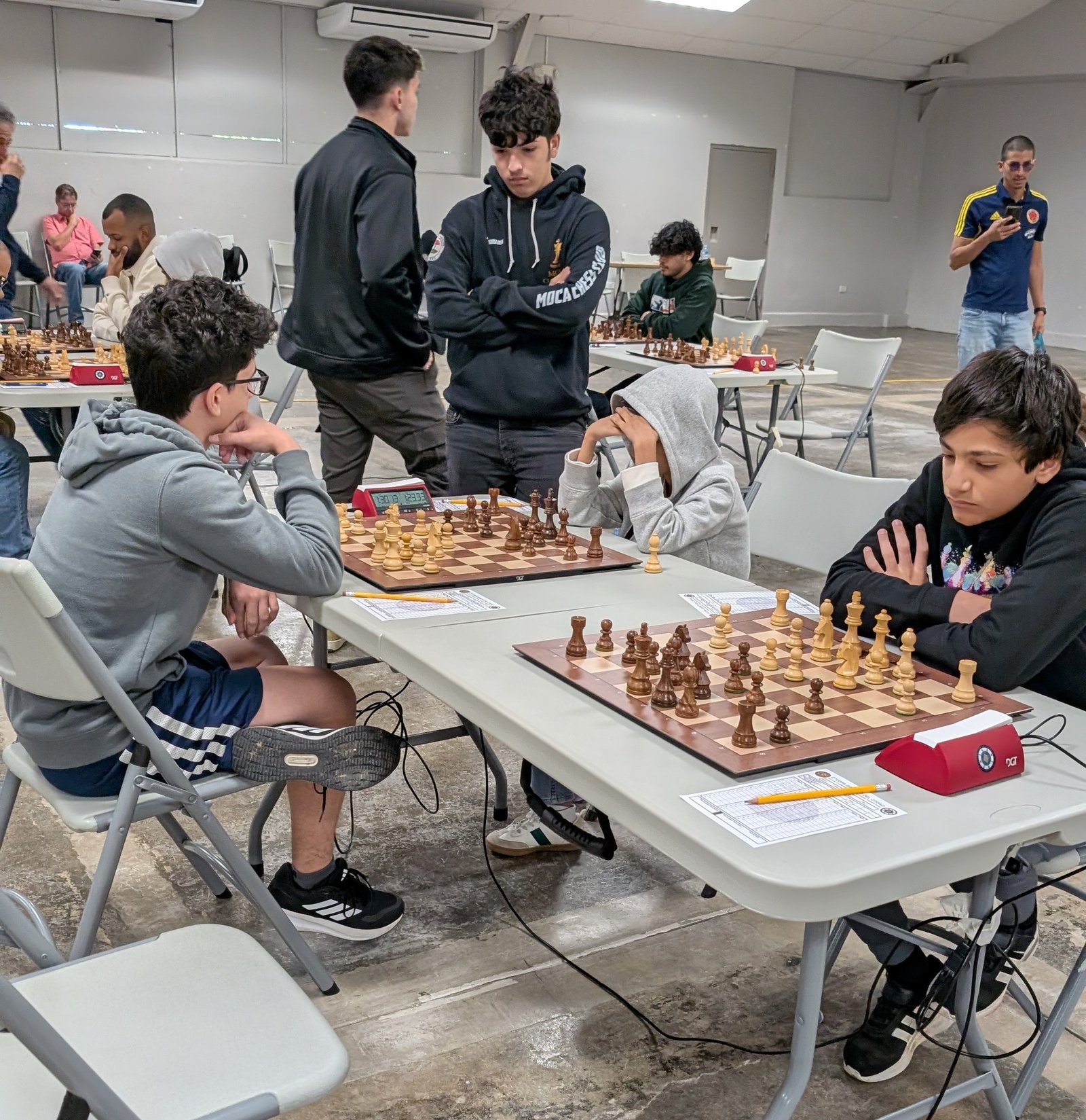 En una misma mesa de la Clase A coinciden Joshua Pedrosa Rivera de pie, Siddhant Tavhare sentado de frente, y de espaldas Stefan Marín Rivera y Christian Rivera Valentín, en la 2da Copa Gambito del Otoao en Utuado.