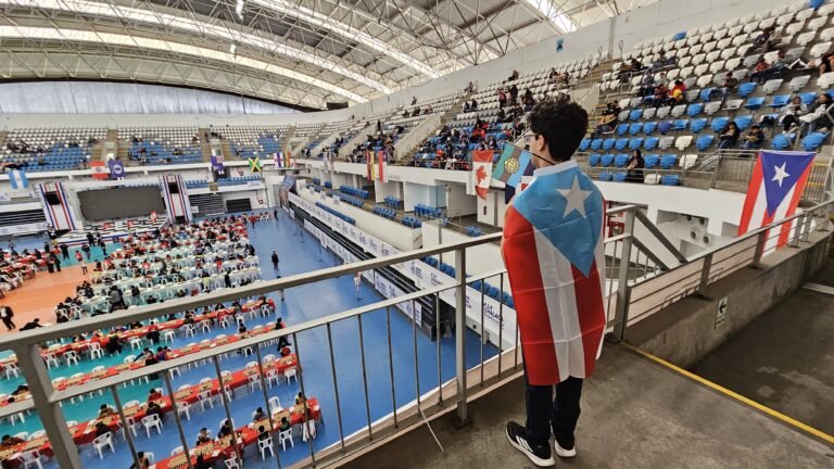 Stefan J. Marín Rivera, envuelto en la bandera de Puerto Rico y sosteniendo la bandera de Utuado, observa desde un balcón el salón de juego del Campeonato Panamericano de la Juventud en Lima.