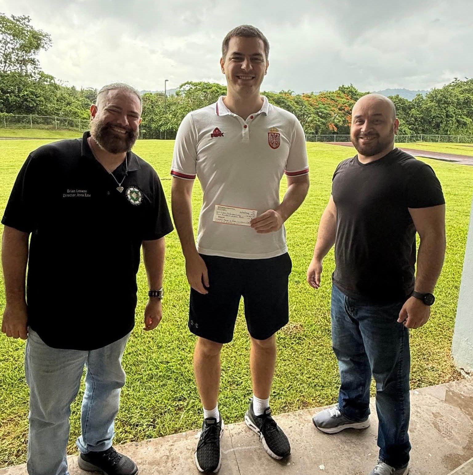 Tres hombres de pie al aire libre en Utuado; el del centro sostiene un cheque de premiación del Torneo de Padres.